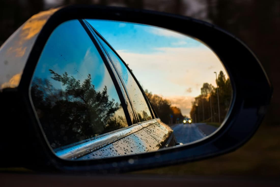 A car’s side mirror reflecting a clean road with a sunset sky, water droplets visible on the mirror and car body.